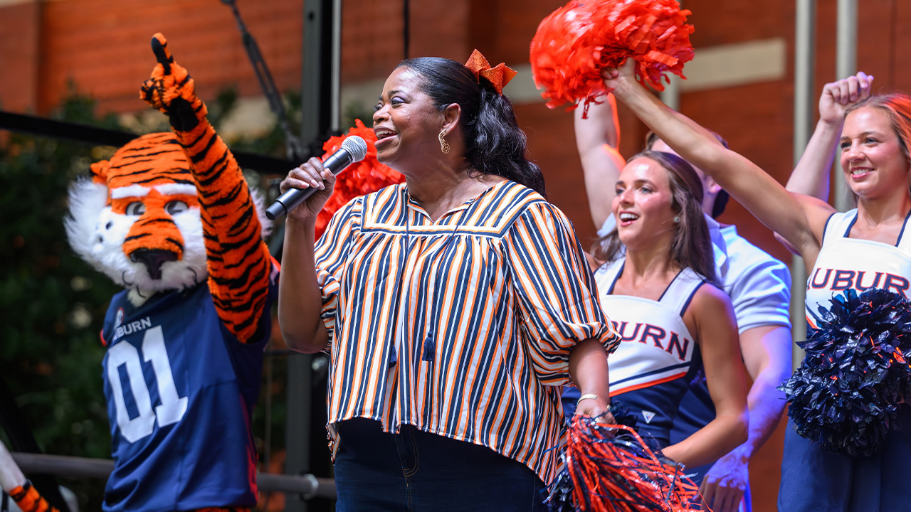 Octavia cheers into a microphone while Aubie and the cheerleaders cheer behind her