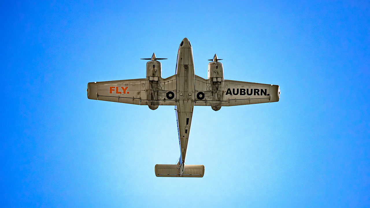 A plane flying overhead with the words "FLY AUBURN" painted on the underside of its wings