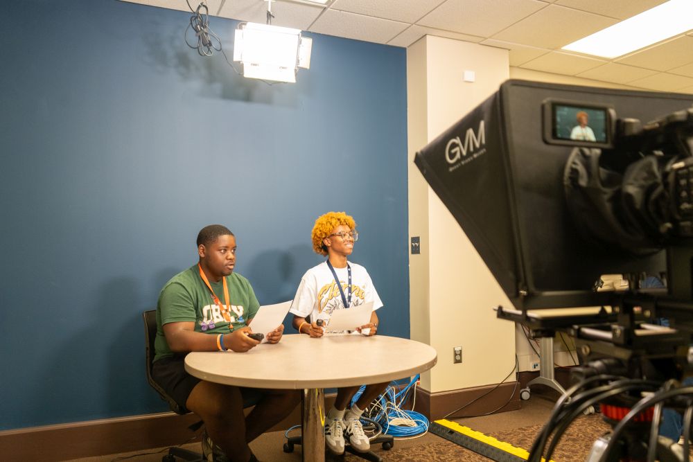 two students sitting at a table in front of a camera pretending to be reporters