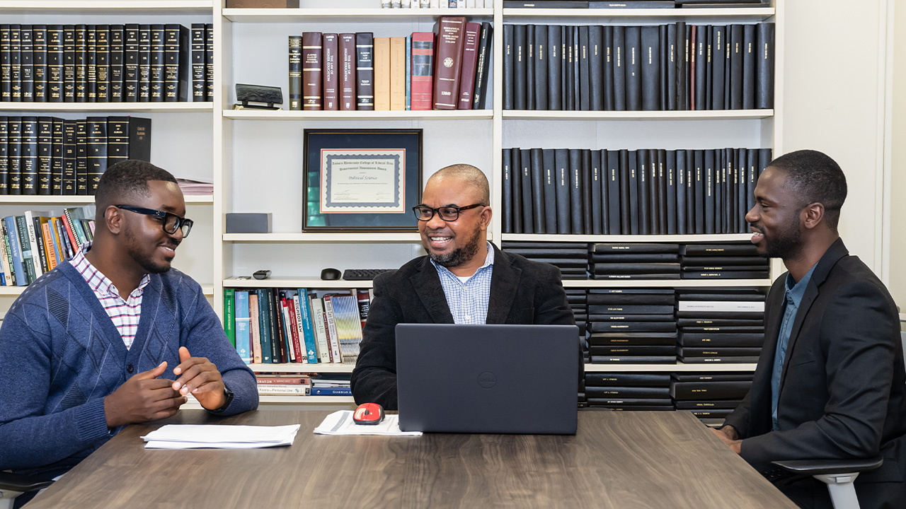 John and two young men sit at a table chatting with bookshelves behind them