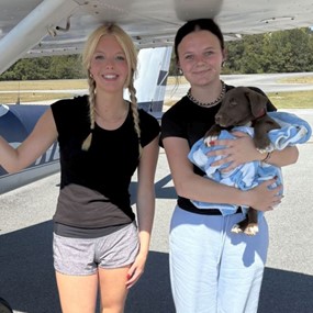 Two women standing under a plane's wing, one is holding a puppy wrapped in small blanket.