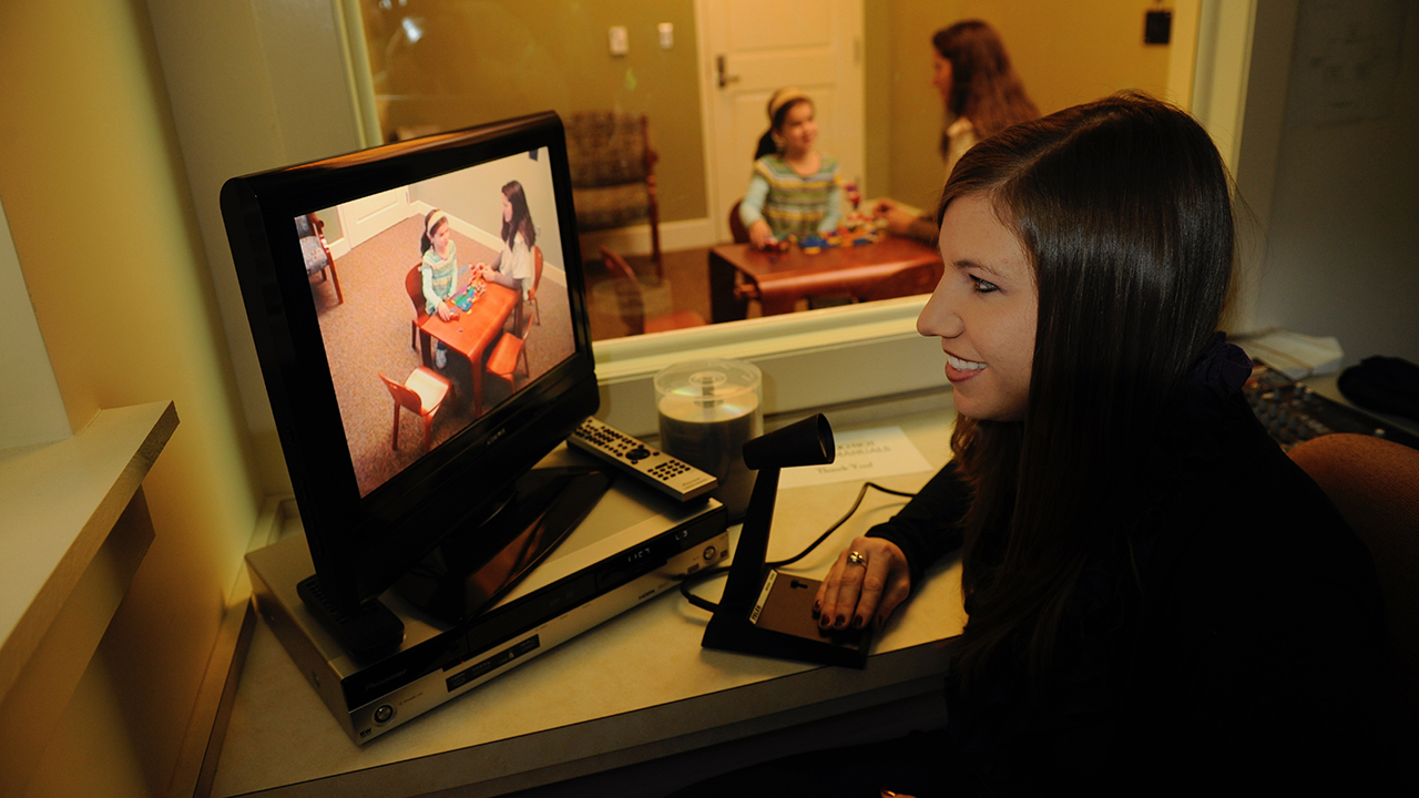 A therapist watches an adult and child build blocks on a TV screen while the same child and adult can be seen through a one-way mirror in the background.