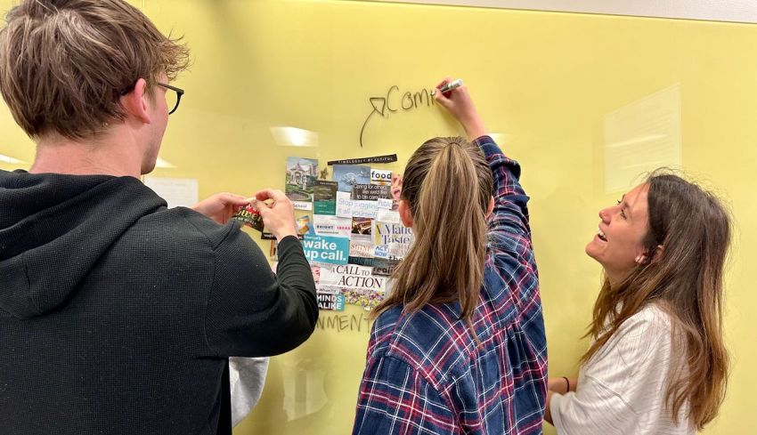 Three students focus on a sticker-covered board, collaborating and exchanging ideas in a lively classroom setting