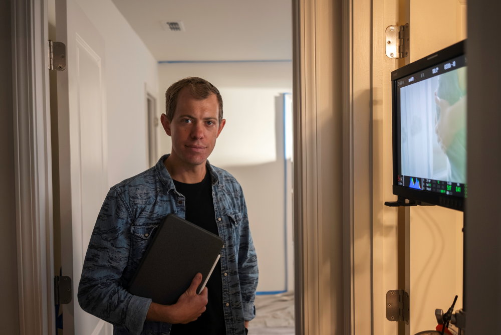 Brock Hanson wearing a denim shirt holds a notebook while standing in a softly lit hallway, looking at the camera.