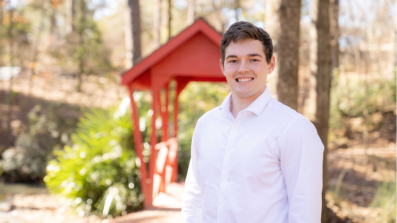 Biomedical sciences sophomore John Walker stands outdoors on Auburn University’s campus near a small red footbridge, wearing a white button-down shirt and smiling toward the camera