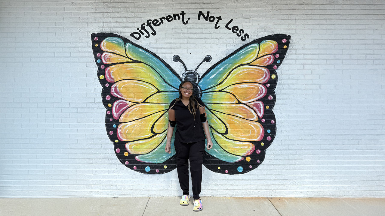 Celeste, who wears prosthetic arms, stands in front of a brick wall painted with butterfly wings