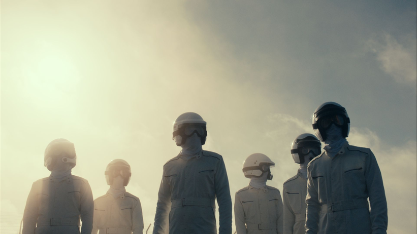 Formula One racers in suits and helmets walking toward the camera with the sun shining behind them