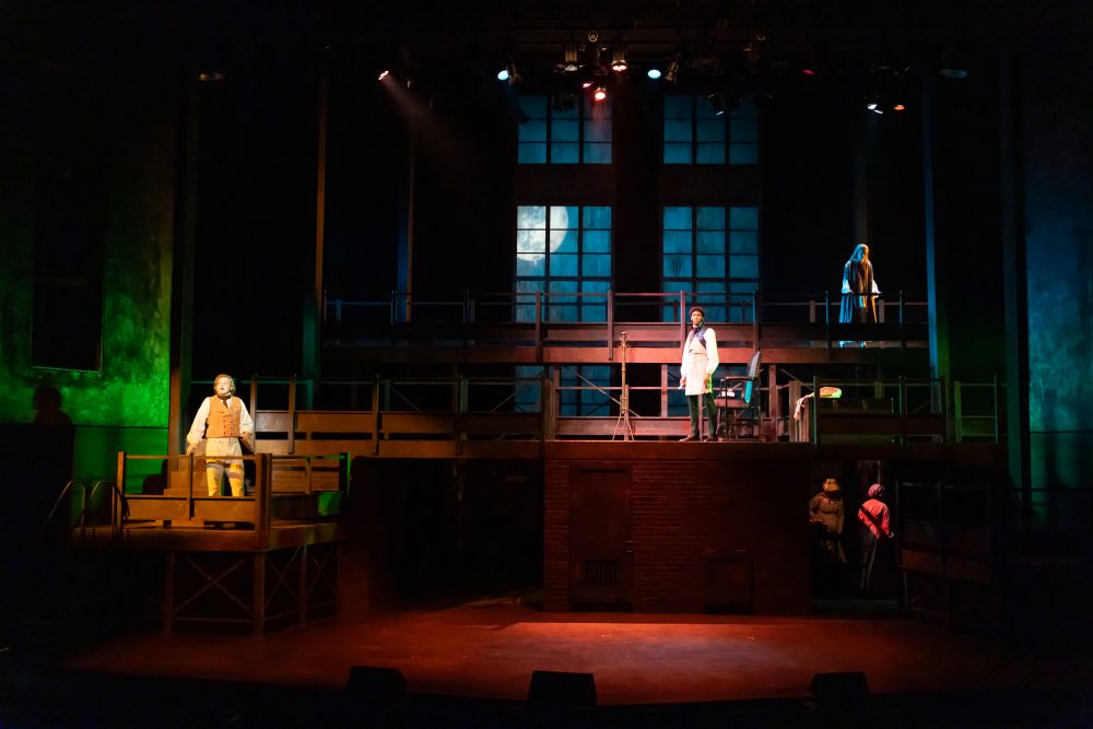 Blue and green lights illuminate specific areas of the stage and a large window projection unit shows a full moon